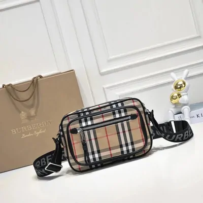 A small rectangular crossbody bag in beige checked canvas with black leather trim and two silver zips, standing on a white table. The wide webbing strap is printed with lettering. A paper carrier bag leans behind it and a white rabbit figurine holding gold spheres sits to the right.