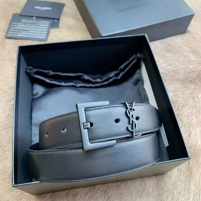 A dark grey leather belt coiled inside its presentation box on a satin dust bag. The buckle is a wide matte rectangle and a metal monogram keeper sits beside it. The box lid and a printed card rest on the fur surface behind.