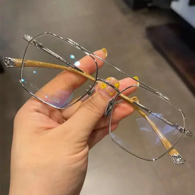 A hand holding a folded pair of silver metal frames with a double bridge and angular lenses. The temples are marbled amber, each hinge and tip carries a small engraved cross, and the clear lenses throw a faint blue reflection.