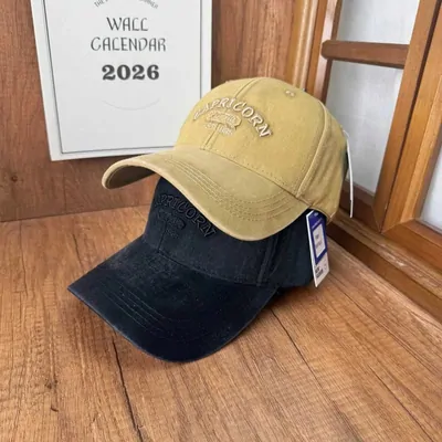 Two washed baseball caps stacked on a wooden floor, a sand coloured one resting on a navy one. Both have a curved brim and arched tonal embroidery across the front panel, and a paper tag still hangs from the upper cap on a string. A framed wall calendar leans behind them.