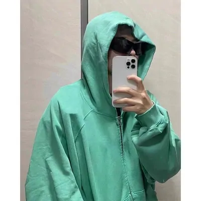 Someone in a mint green zip-through hoodie taking a mirror photo, hood pulled up over a cap, dark sunglasses on, phone raised in front of the face. The cut is oversized with dropped shoulders and the zip is open at the chest.
