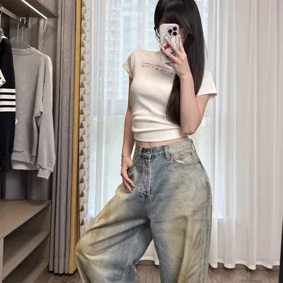 A bedroom mirror photo. A cropped fitted top above wide leg jeans in a faded sandy blue wash, high at the waist and pooling at the hem. A rail of hanging clothes stands to the left and daylight comes through sheer curtains.