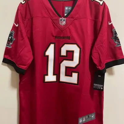 A red American football shirt hanging flat against a plain wall. A large white player number outlined in black and orange fills the chest, an arched team name sits above it, a league shield is stitched at the neck, and a pirate ship patch is sewn to each sleeve. A dark tag hangs at the side seam.
