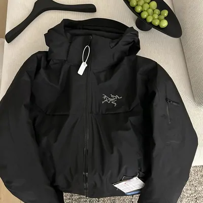 A black hooded shell jacket laid flat on a pale sofa, zipped most of the way up, with a small embroidered motif on the chest, a zipped sleeve pocket, a blank hang tag at the collar and a printed care card near the hem. A hanger and a dish of green grapes lie beside it.