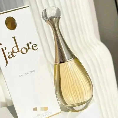 An amphora shaped bottle of pale gold liquid with a ribbed gold neck and a frosted round stopper, leaning against its white carton. The carton is printed in gold script and has a gold window near the top. Ribbed white fabric falls behind.
