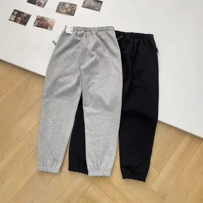 Two pairs of fleece joggers laid side by side on a wood floor, one marl grey and one black. Both have a drawstring elastic waist and a gathered cuff at the ankle. Small photo prints are taped to the wall above them.