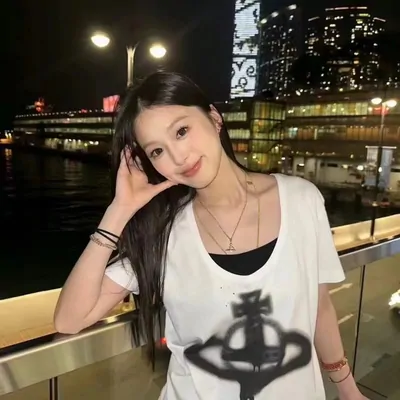 A woman leaning on a waterfront railing at night, wearing a loose white tee over a black top with a sprayed dark motif across the front. Lit towers and harbour water sit behind her.