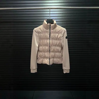A short fitted jacket on a hanger against a dark slatted shutter. The front panel is quilted in horizontal channels, the sleeves and sides are plain knit, the collar stands upright, and a small oval patch sits on one arm. The colour is a pale blush.